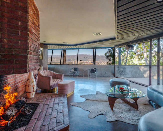 A modern living room features a curved sofa, a glass coffee table, and a fireplace, with large windows offering a view of the surrounding landscape.