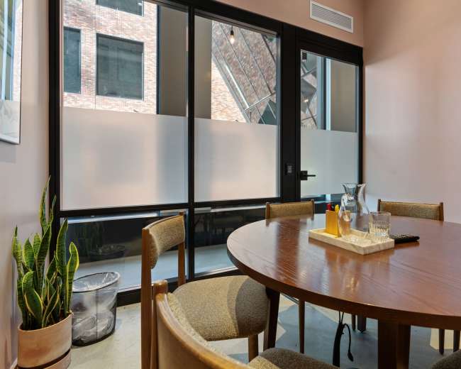 A wooden conference table is set with a pitcher and glasses, while a large window offers a view of the exterior brick wall.