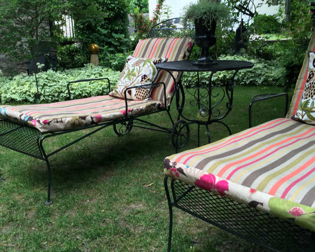 Two striped lounge chairs with decorative cushions sit beside a small metal table in a grassy garden surrounded by greenery.