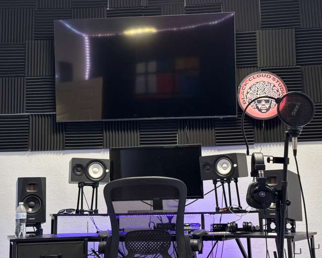 The image shows a music studio setup with a large wall-mounted TV above a black desk featuring audio equipment, monitors, and a microphone.