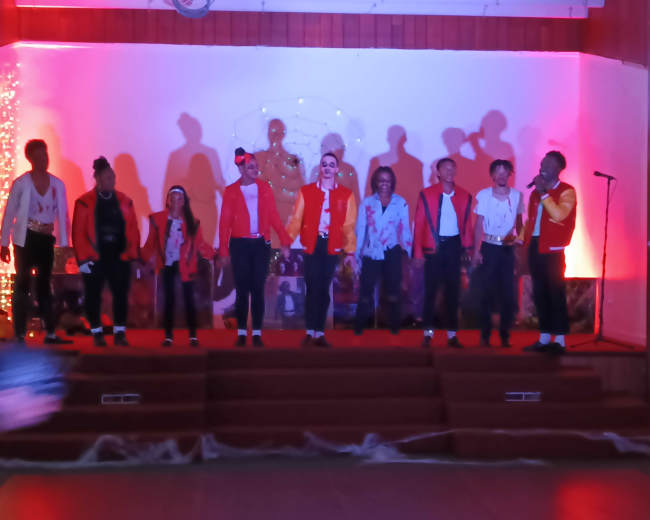 A group of performers in matching jackets stands on a stage with colorful lights and decorations.