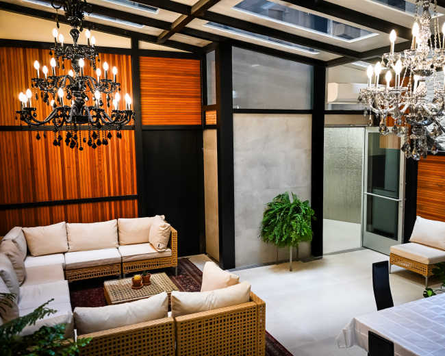 The image shows a modern indoor lounge area featuring wicker seating, chandeliers, and a glass ceiling with wooden paneling.