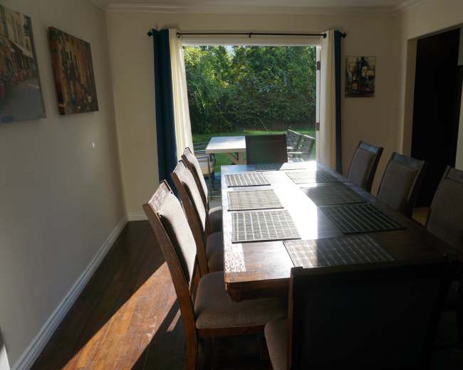 A wooden dining table with eight chairs is set against a window that overlooks a green backyard.