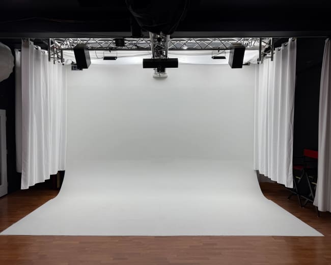 The image shows a photography studio with a white backdrop, soft lighting, and curtain drapes on either side.