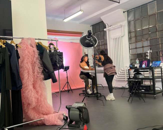 A photography studio setup features a model sitting at a table while a stylist adjusts her makeup, surrounded by clothing racks and a backdrop.