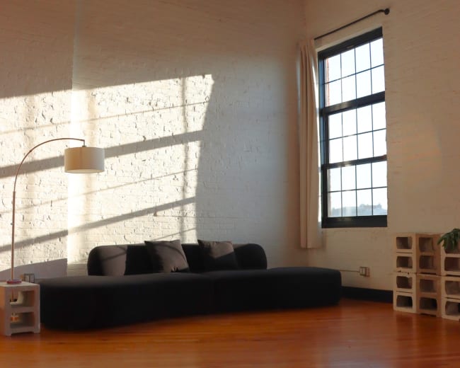 The image shows a modern living space with a sectional sofa, a floor lamp, a large window, and a patterned rug on wooden flooring.