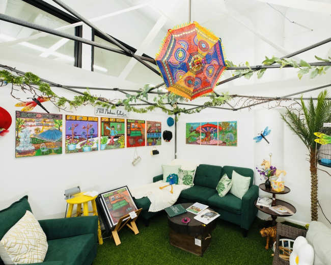 The image shows a cozy indoor space featuring green sofas, colorful wall art, and decorative plants, with a vibrant chandelier hanging from the ceiling.