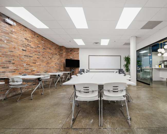 The image shows a modern conference room featuring a large white table surrounded by several white chairs, with exposed brick walls and a glass partition.