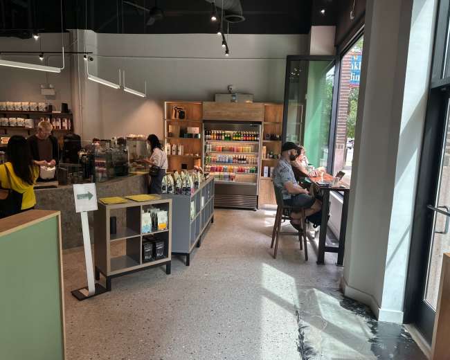 The image shows the interior of a café with a coffee counter, shelves lined with beverages, and customers seated at tables near large windows.