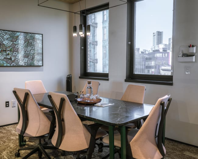 A modern conference room features a large table surrounded by six chairs, with a decorative painting on the wall and windows displaying a cityscape.