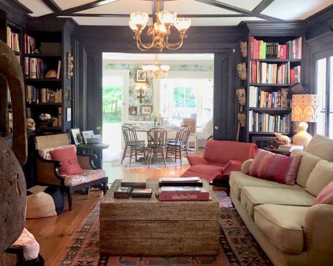 A cozy living room with dark wood accents, a chandelier, a large sofa, and bookshelves filled with books, leading to a bright dining area with a table and chairs visible through large windows.