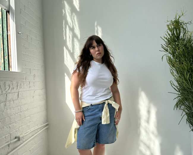 A person stands in a brightly lit room with white walls and a large plant, dressed in a white top, denim shorts, and yellow shoes.