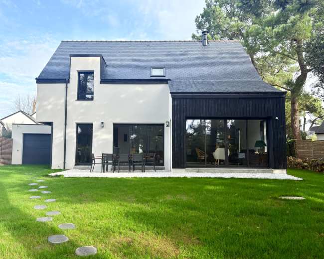 The image shows a modern house with a blend of white walls and a black wooden facade, surrounded by a well-maintained lawn and a stone pathway.