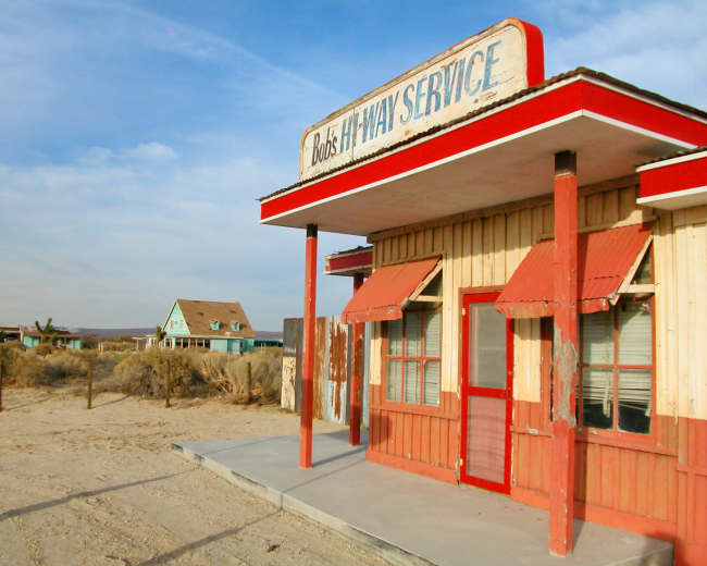 A weathered service station building with a red trim, labeled "Bob's Highway Service," standing on a sandy lot near an abandoned house.