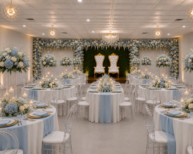 The image depicts an elegantly decorated banquet hall featuring round tables set with blue and white floral centerpieces and two ornate white chairs positioned on a stage at the back, surrounded by flower arrangements.