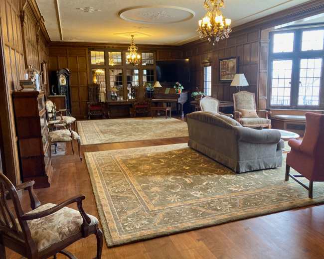 The image shows a spacious, elegantly furnished room with wooden paneling, a large area rug, and multiple light fixtures.