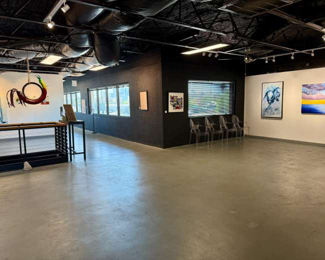 The image shows an art gallery with a spacious, open floor layout featuring various artworks on the walls and transparent chairs arranged in a seating area.