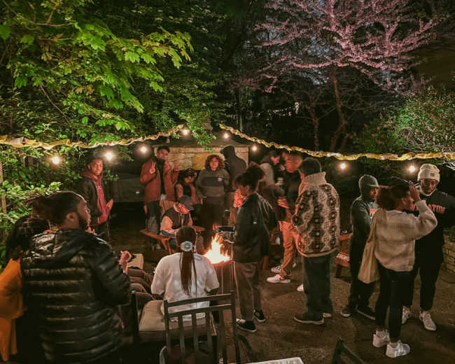 A group of people gathers around a fire in a well-lit outdoor setting surrounded by trees and decorative lights.