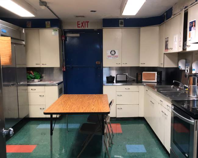 A kitchen with a central table and two chairs, surrounded by cabinets, appliances, and an exit door.