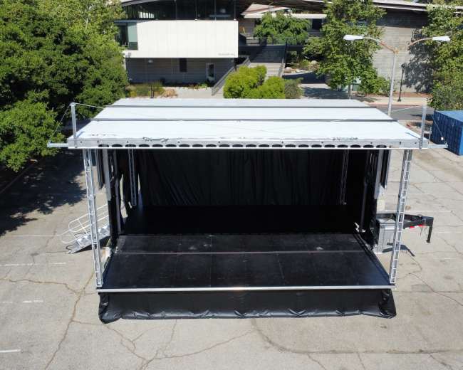 An empty outdoor stage with a black backdrop is set up in a parking lot surrounded by trees and buildings.