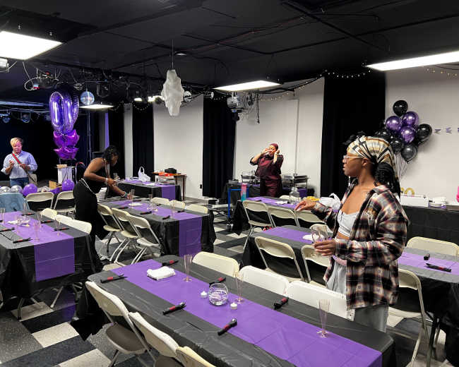 A party setup in a decorated room with purple tables and chairs, a stage area, and balloons, while several people are preparing for the event.