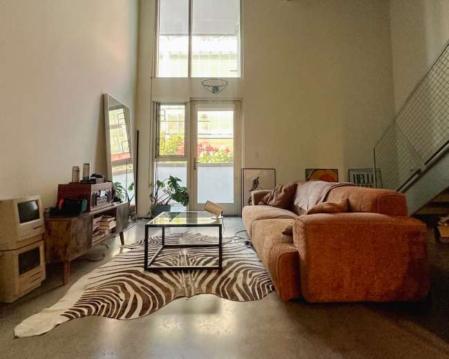 A spacious living area features a large orange couch, a glass coffee table on a zebra-print rug, and a mirror against a wall with large windows allowing natural light.