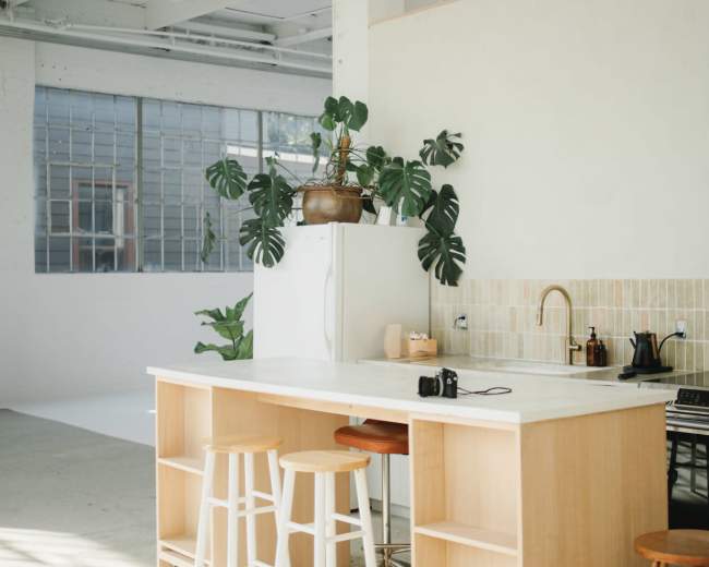 The image features a modern kitchen with a light wood island, two stools, plants, and a minimalist design.