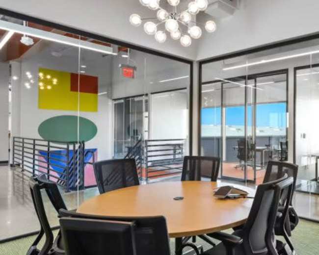 The image shows a modern office space with a round conference table surrounded by black chairs, and large glass panels separating different work areas.