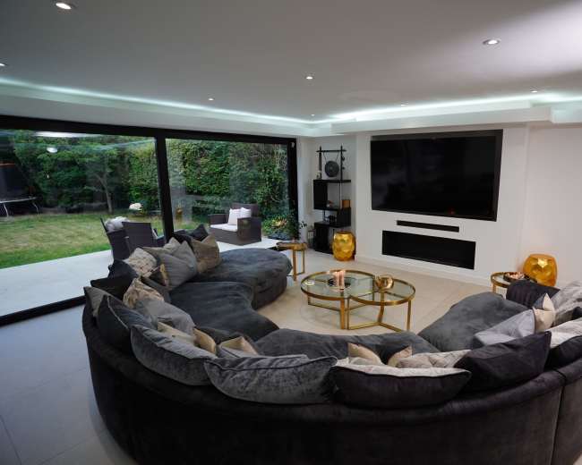 The image shows a modern living room featuring a large curved gray sofa, a glass coffee table, and a wall-mounted television, with sliding glass doors leading to a green garden outside.