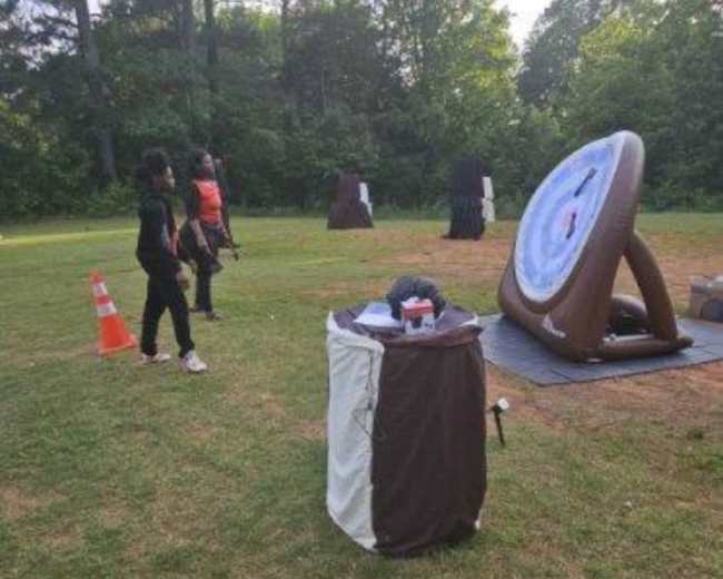 The image shows a grassy area with a large archery target, cones, and several individuals preparing to shoot arrows.