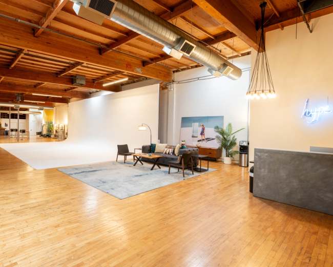 The image showcases a spacious, modern interior with wooden floors, exposed beams, and a large white backdrop area, complete with seating and decorative elements.