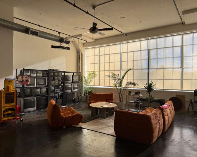 The scene showcases a spacious, well-lit room with large windows, featuring a collection of vintage televisions, a round coffee table, and cozy orange lounge furniture surrounded by potted plants.