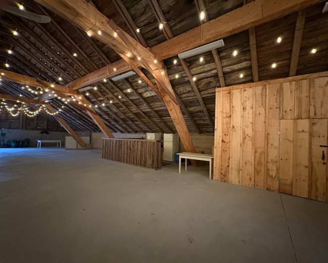 The image shows a spacious attic with wooden beams and string lights, featuring a wooden bar area and a storage closet in one corner.