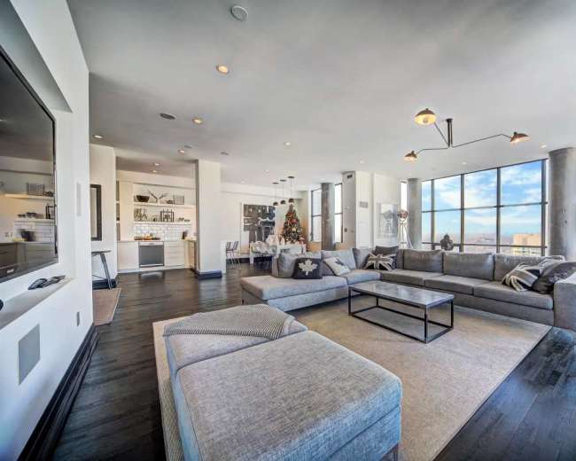 The image shows a modern living room with large windows, a sectional sofa, and a coffee table in a high-rise apartment.