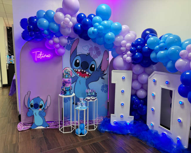The image features a colorful birthday party setup with a large "10" made of white balloons, a decorated table holding various treats, and a backdrop of blue and purple balloons alongside a character illustration.