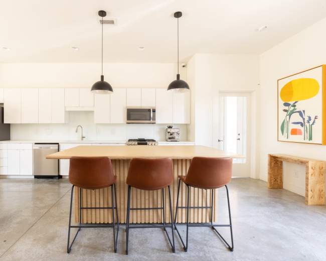 The image shows a modern kitchen with white cabinetry, stainless steel appliances, and a central wooden island with three brown bar stools.