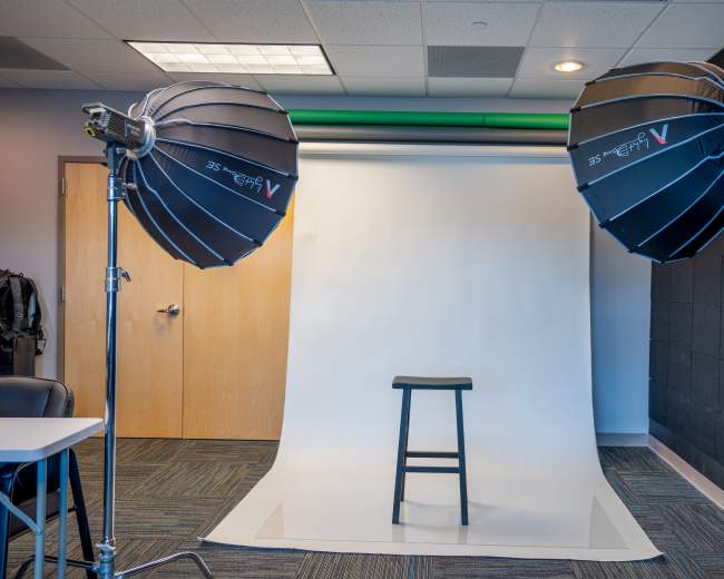 The image shows a photography studio setup featuring two softbox lights positioned on either side of a stool placed in front of a white backdrop.