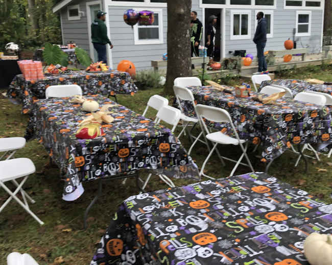 A backyard decorated for a Halloween party features tables with themed tablecloths, pumpkins, and guests socializing near a small gray house.