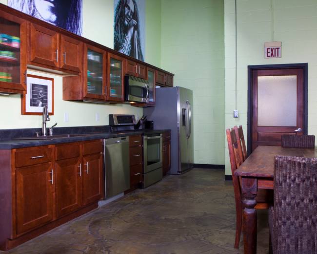 The image shows a kitchen with wooden cabinets, stainless steel appliances, and a dining table made of wood, complemented by wicker chairs.