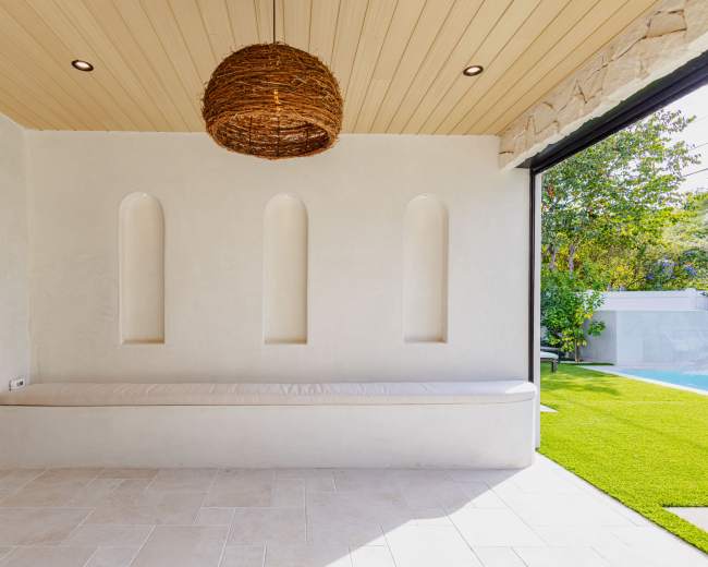 The image shows a bright indoor space featuring a smooth white wall with three recessed niches and a cushioned bench beneath a woven light fixture.