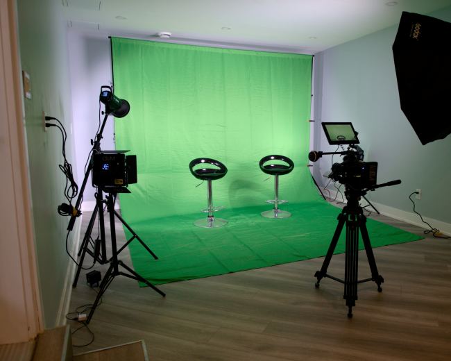 A photography studio with two black chairs on a green backdrop, illuminated by various lights and equipped with a camera on a tripod.