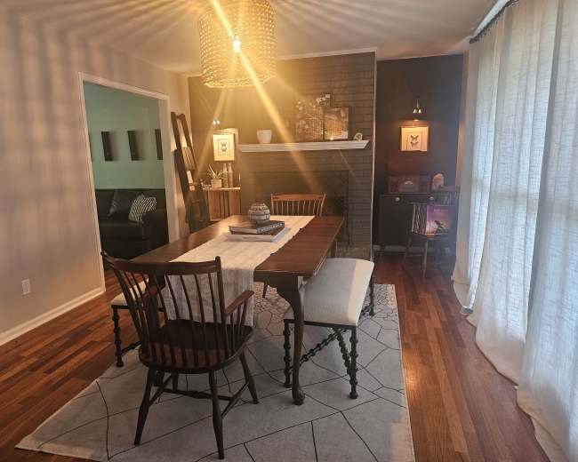 The image shows a dining area featuring a wooden table with chairs, a patterned rug, a fireplace, and large windows draped with light-colored curtains.