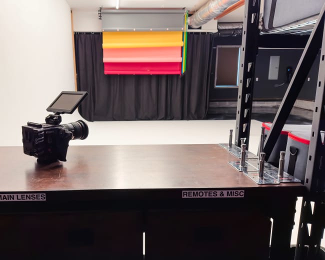 A camera is positioned on a wooden table labeled "MAIN LENSES," facing a backdrop with colorful horizontal panels in a studio setting.