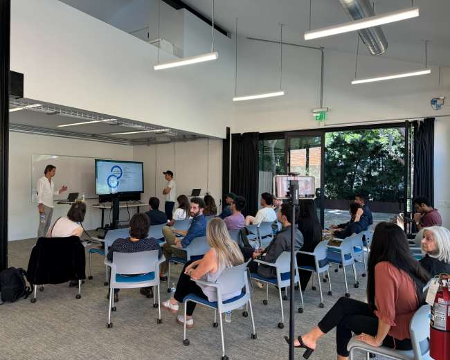 A group of people sits attentively in a classroom setting, watching a presentation given by a speaker at the front of the room.