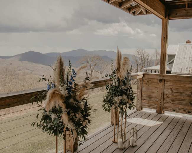 Two floral arrangements with blue flowers and pampas grass stand on a wooden deck overlooking a mountainous landscape under a cloudy sky.