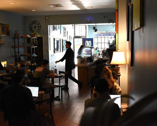 A man in a suit walks through a cafe where several customers are seated, some working on laptops.