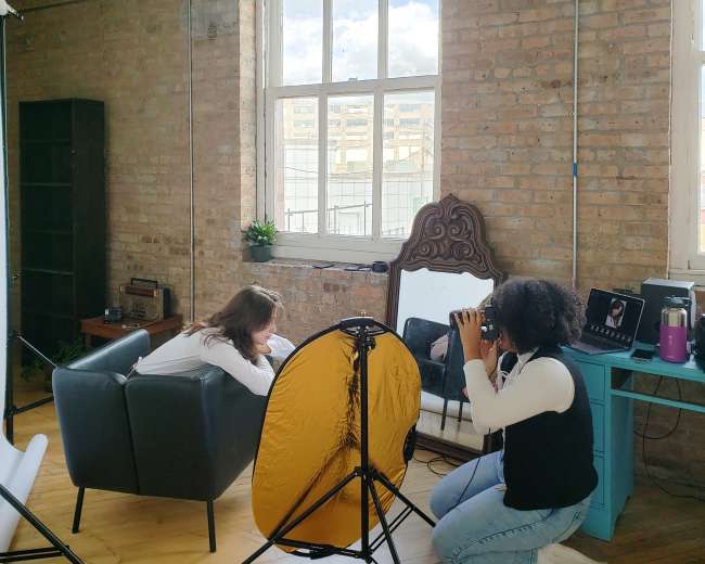 A photographer captures an image of a model sitting on a sofa in a studio setting with large windows and a backdrop.