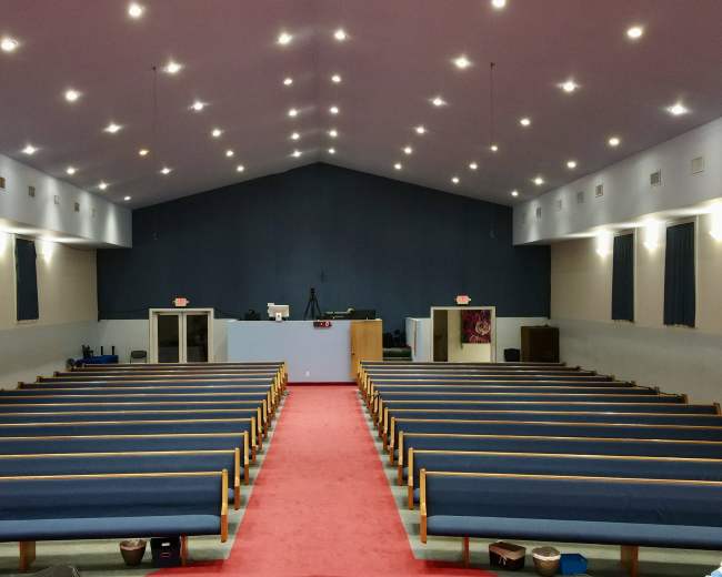 The interior of a church features rows of blue pews facing a stage area at the front, with a red carpet running down the center and overhead lights illuminating the space.