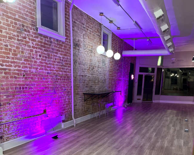 A spacious interior with a wooden floor, exposed brick walls illuminated by purple lighting, and minimal furniture.
