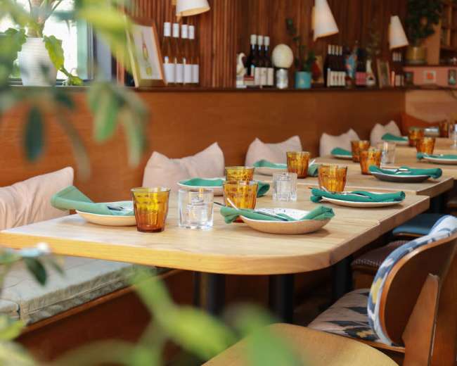 The image shows a cozy restaurant interior with wooden paneling, a wooden dining table set with green napkins, amber glassware, and cushioned seating.